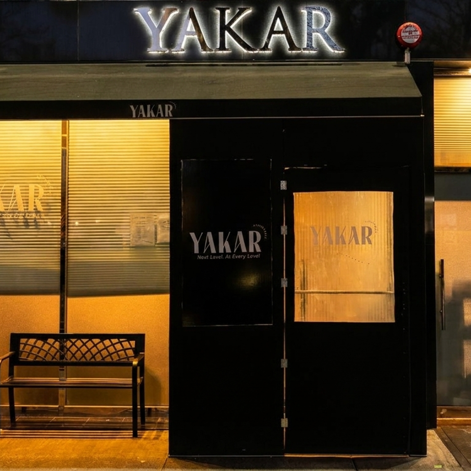 Nighttime street view of Yakar restaurant entrance, featuring illuminated signage, black doors, amber windows, and a bench.